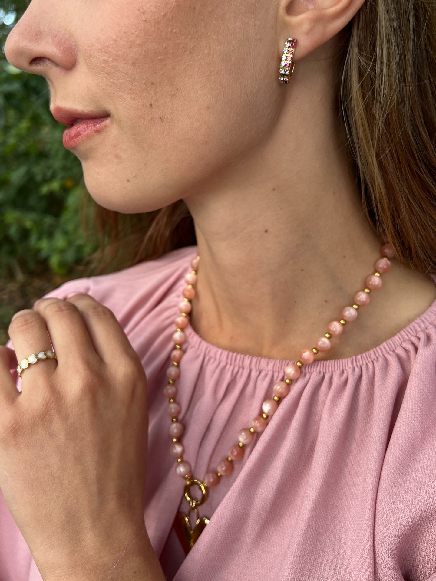 Close-up van vrouw met roze top, oorbellen en kralenketting, buiten bij groen, zijaanzicht.