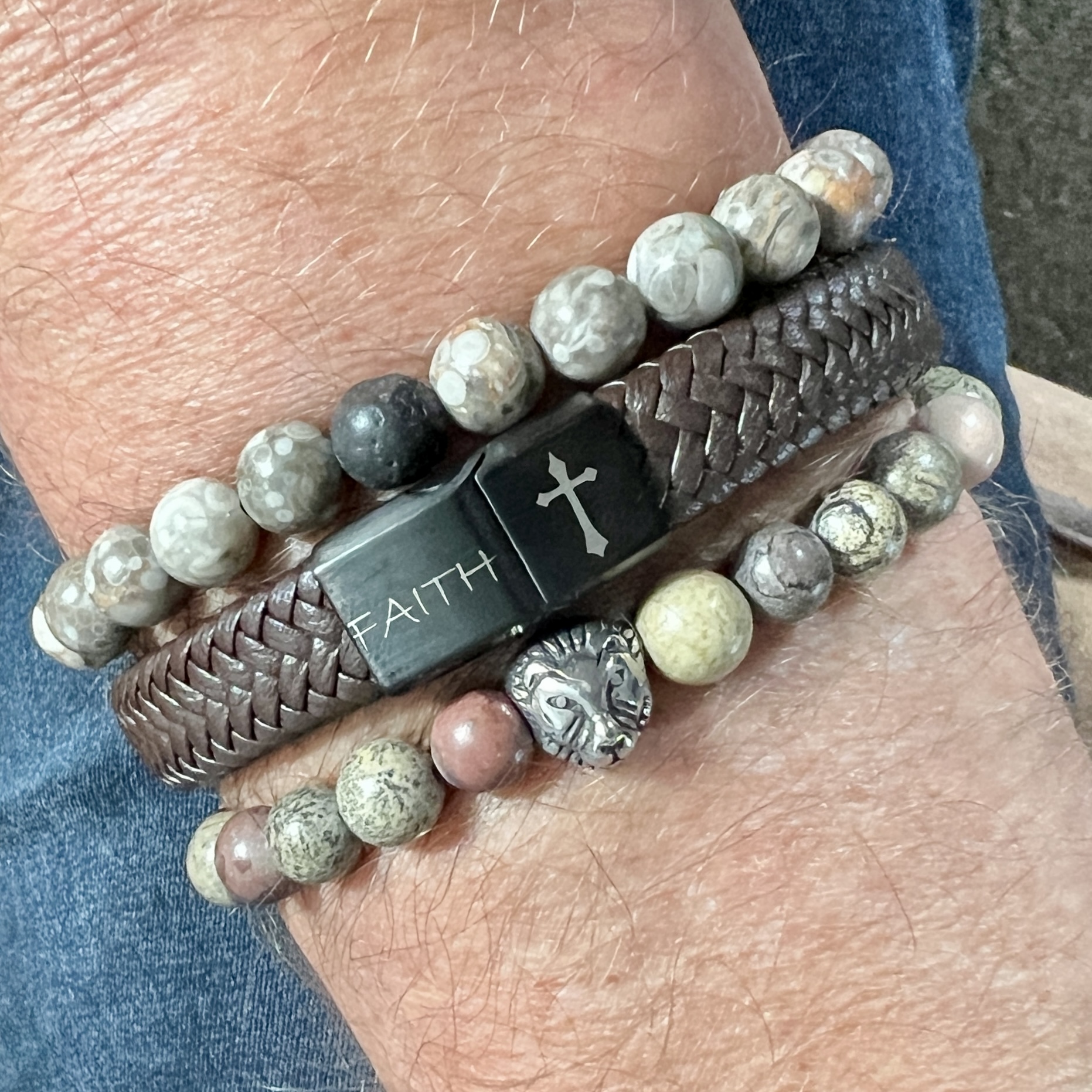 Hand met kralenarmband van grijze bruine stenen, zilveren leeuwenkop kraal en bruin leren armband met zwart slotje met witte tekens faith en een kruisje