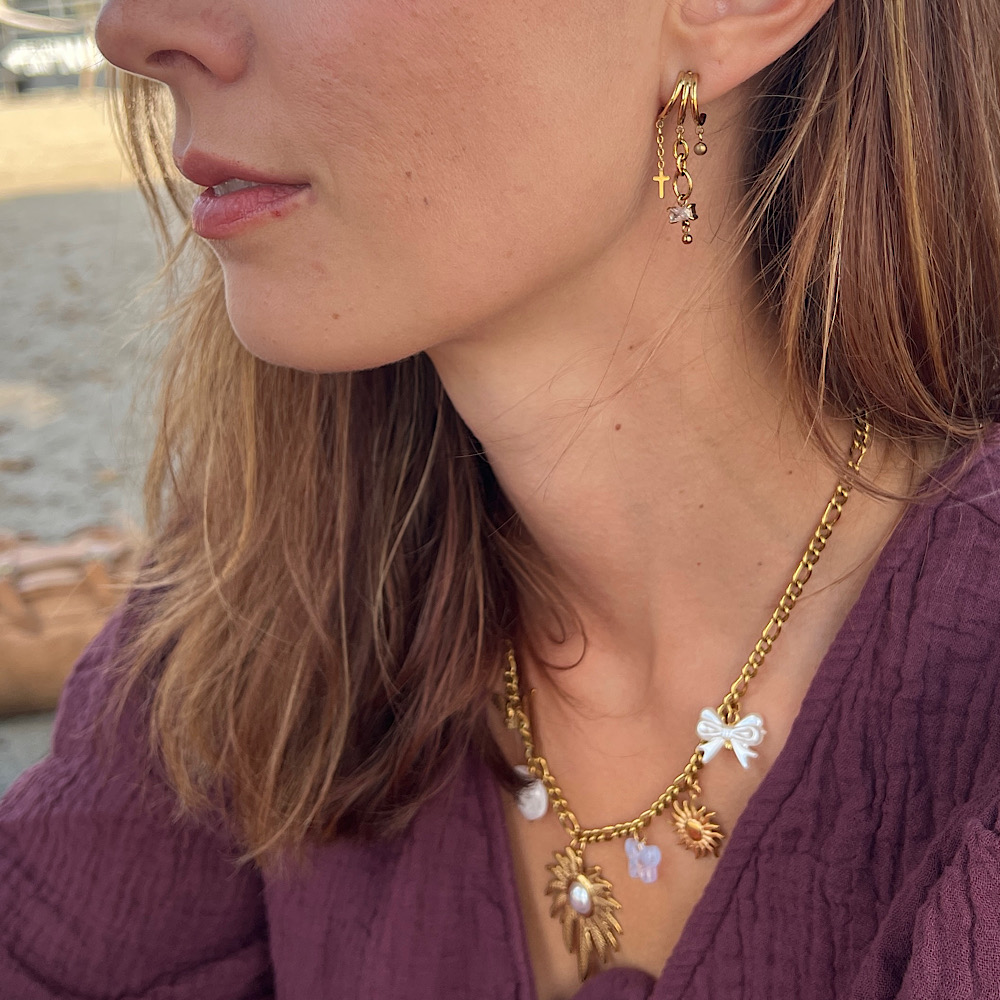 Close-up van vrouw met gouden oorbellen en kettingen met vlinder- en kruisbedels, in paarse blouse.
