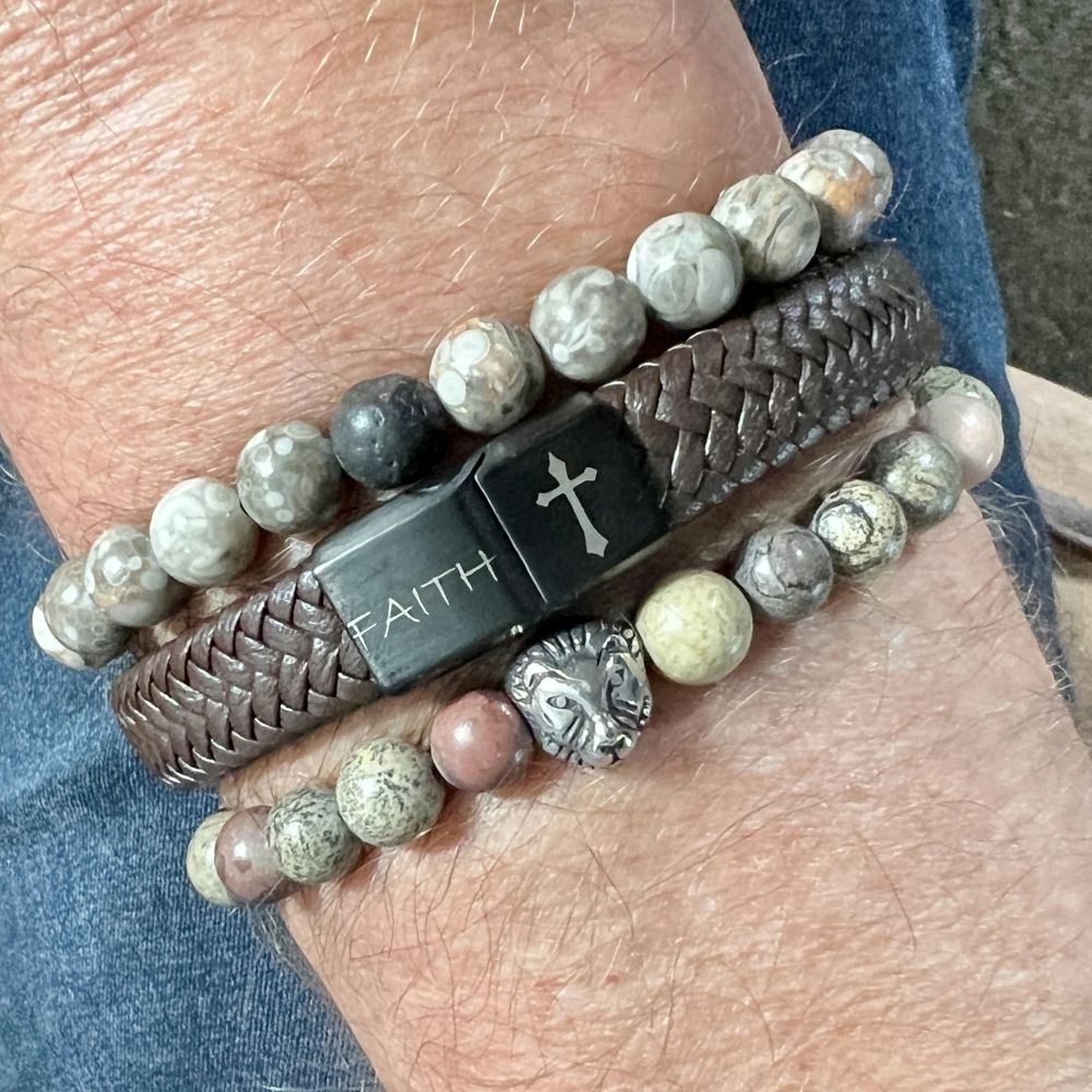 Hand met kralenarmband van grijze bruine stenen, zilveren leeuwenkop kraal en bruin leren armband met zwart slotje met witte tekens faith en een kruisje