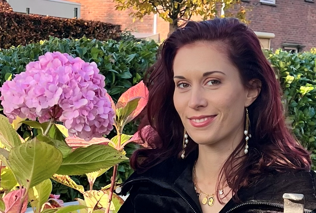 Glimlachende vrouw poseert naast roze hortensia, met bedelketting en lange oorbellen, in een tuin, met bakstenen muur op de achtergrond.