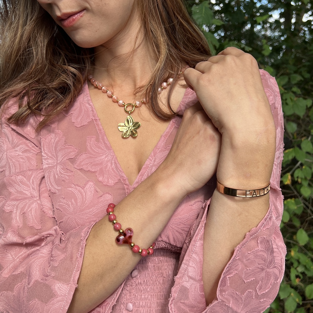 Vrouw draagt roze blouse met gouden ketting en armbanden, hand bij hals, buiten tussen groene bladeren.