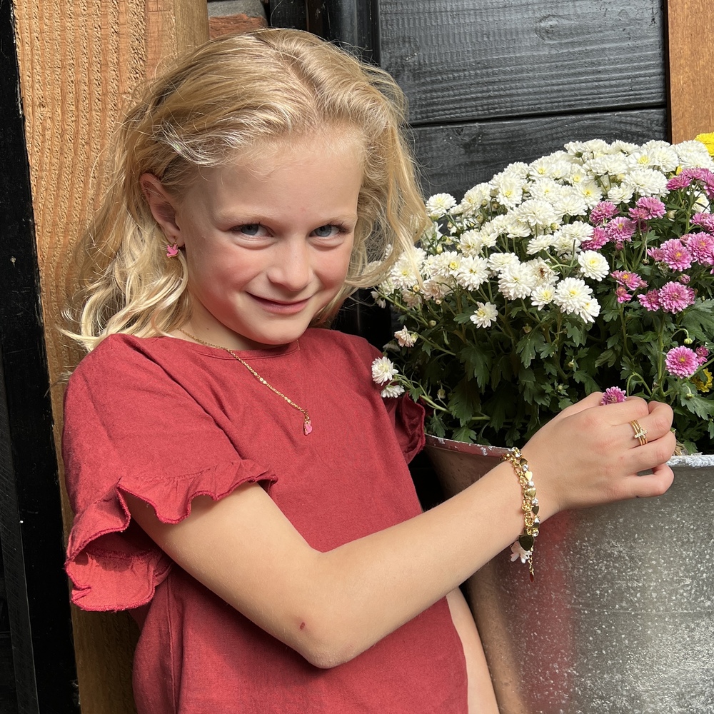 Blond meisje in rode jurk poseert bij bloemenbak met witte en roze bloemen, glimlachend en arm uitgestrekt. Ze draagt sieraden van Grace And Praise