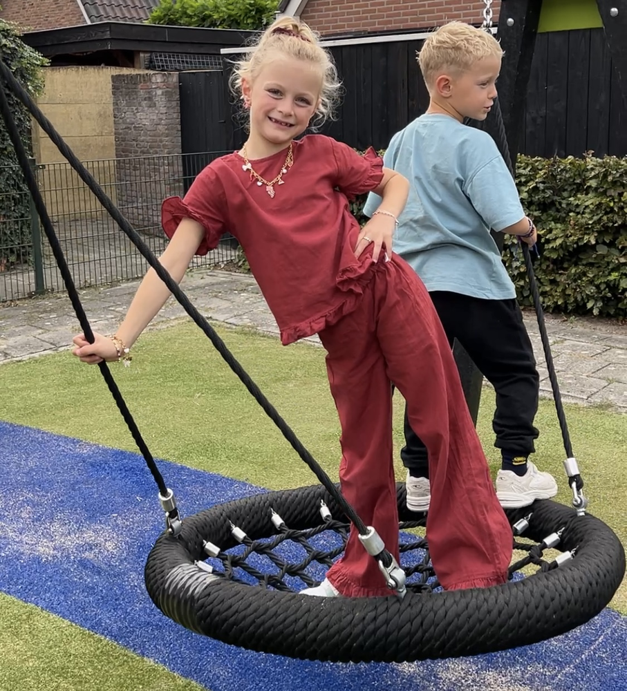Twee kinderen spelen op een band schommel met touwrand in een tuin, meisje in rood, jongen in blauw.