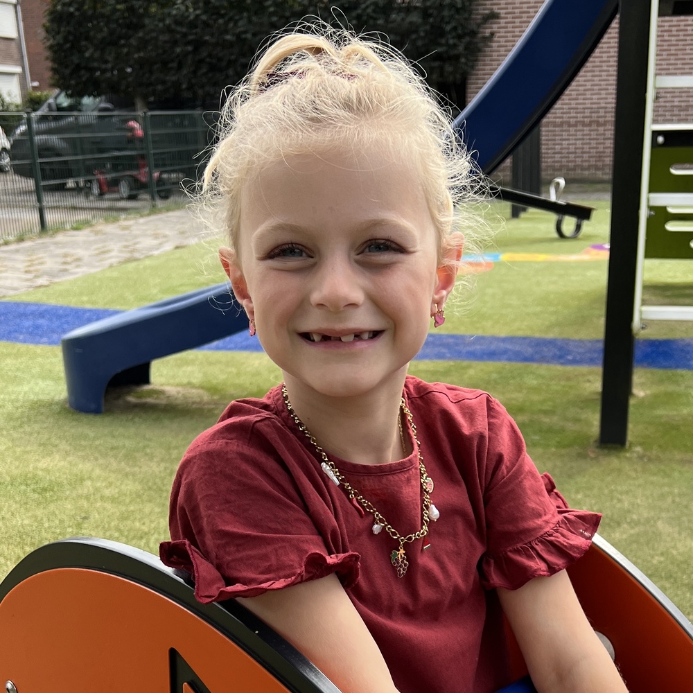 Blond meisje glimlacht op een speelplaats, zittend op een oranje wip-kip, met rode jurk en ketting van Grace And Praise