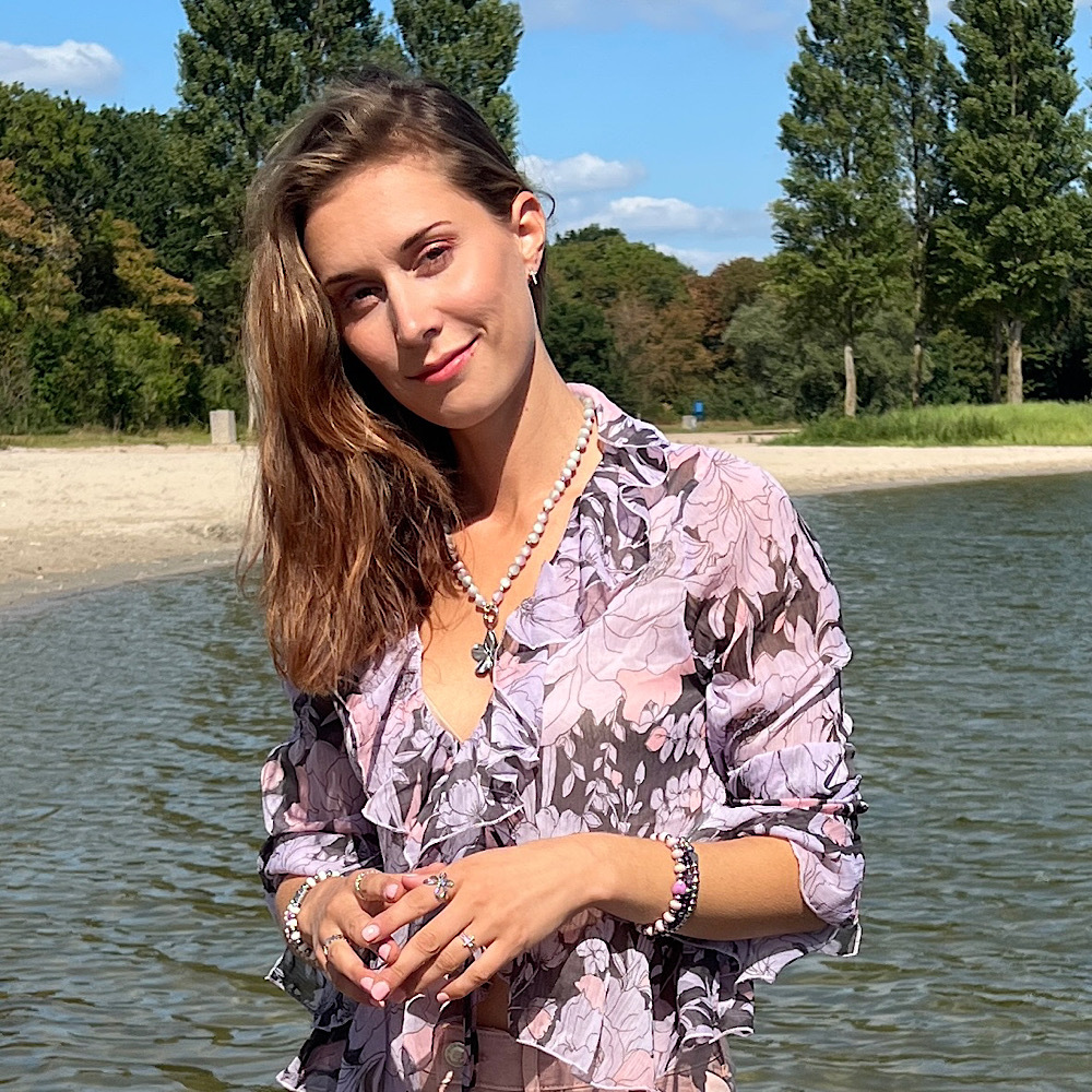 Vrouw met sieraden van Grace And Praise en bloemenjurk poseert glimlachend aan een meer, met bomen en blauwe lucht op achtergrond.