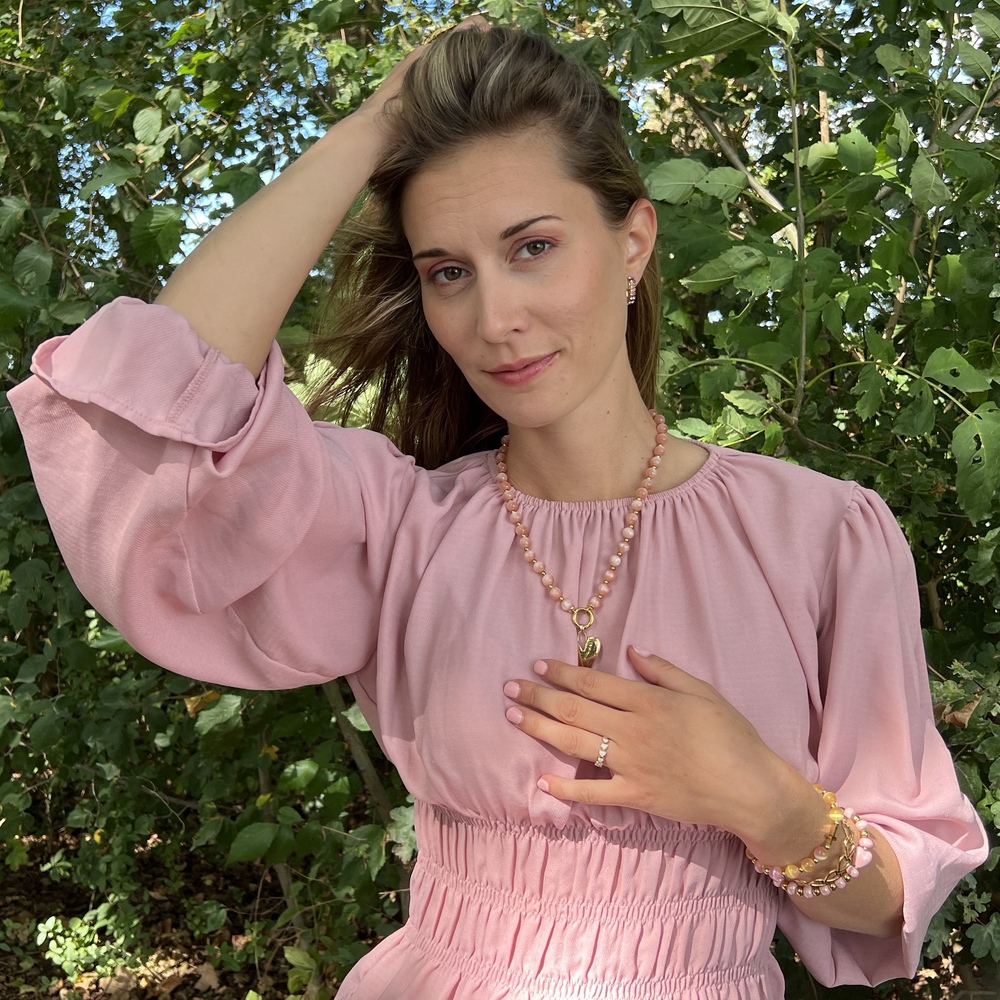 Vrouw met sieraden van Grace And Praise en bloemenjurk poseert glimlachend aan een meer, met bomen en blauwe lucht op achtergrond.